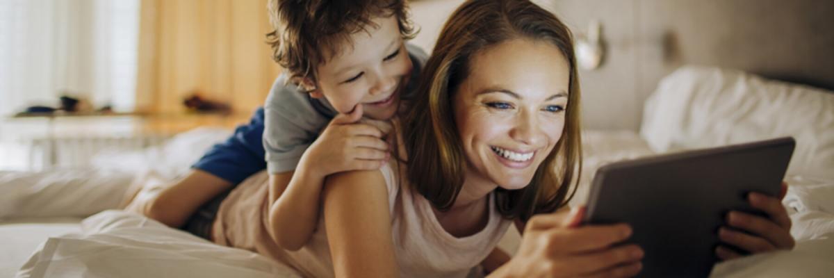 Mother and young son laying on bed smiling with tablet, Make an Appointment